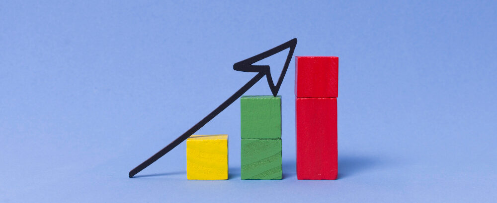 Three wooden blocks stacked in ascending order, with a black arrow pointing upward above them. The blocks are colored yellow, green, and red, representing growth and progress.