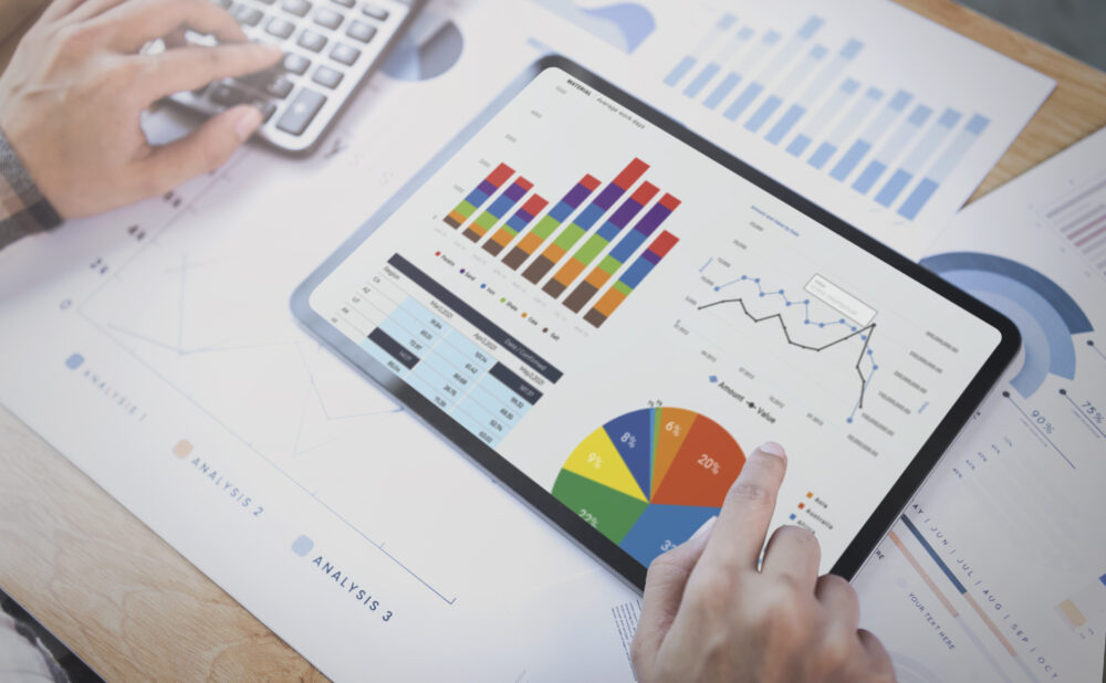 A person is reviewing data on a tablet. The tablet screen displays various charts and graphs, including a bar chart, a line graph, and a pie chart. The person's finger is pointing at one of the graphs.