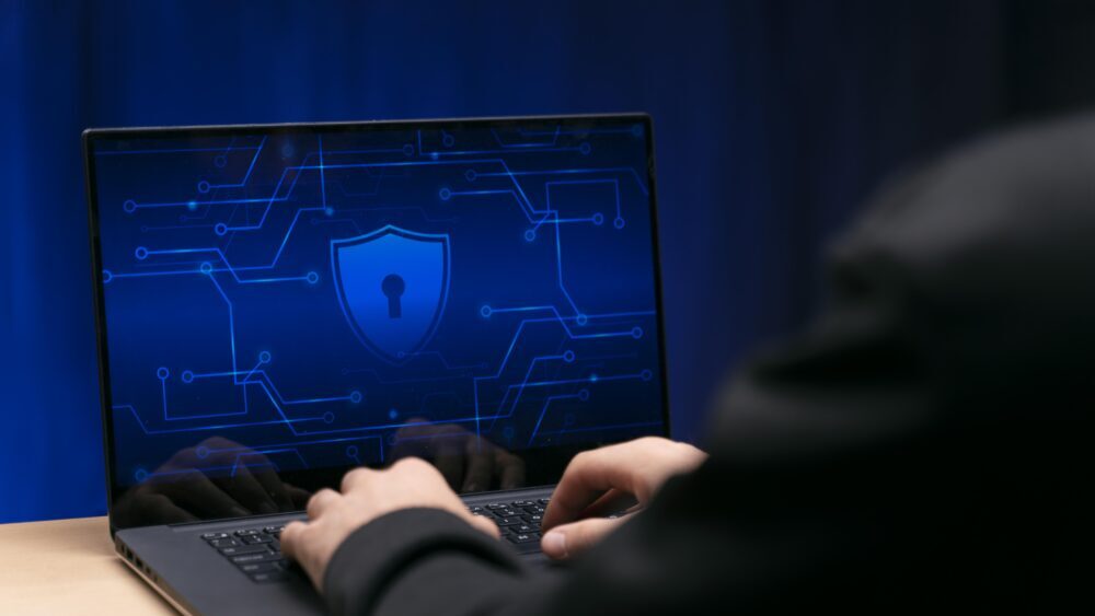 A programmer typing in a laptop, displaying a blue shield in the center of the display.